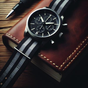 May include: A black chronograph wristwatch with a black leather band and white stripes. The watch is on a brown leather surface.