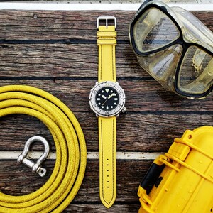 May include: A yellow watch with a black face and a silver bezel and buckle. The watch is on a yellow strap. A yellow diving hose, a silver shackle, a diving mask, and a yellow case are also visible on a wooden surface.
