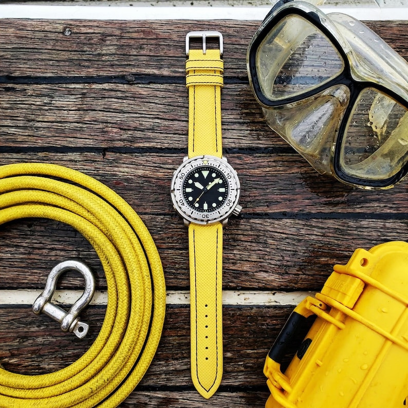May include: A yellow watch with a black face and a silver bezel and buckle. The watch is on a yellow strap. A yellow diving hose, a silver shackle, a diving mask, and a yellow case are also visible on a wooden surface.