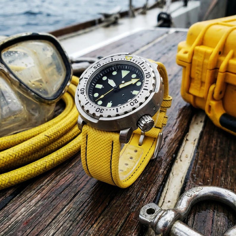 May include: A stainless steel dive watch with a black face and luminous markers, paired with a bright yellow canvas strap. The watch is set against a backdrop of diving equipment, including a mask and a yellow case, suggesting a nautical theme.