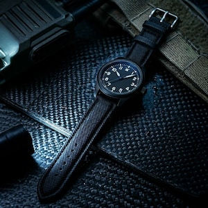 May include: A black wristwatch with a black dial and white numerals. The watch has a black strap with a textured pattern and a silver buckle. The watch is set against a dark, textured background.