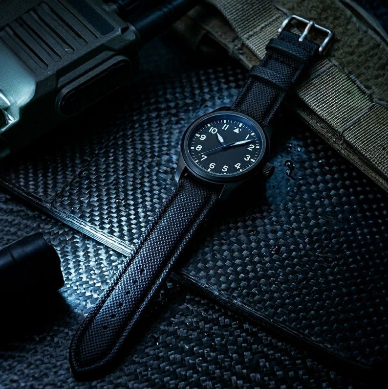 May include: A black wristwatch with a black dial and white numerals. The watch has a black strap with a textured pattern and a silver buckle. The watch is set against a dark, textured background.
