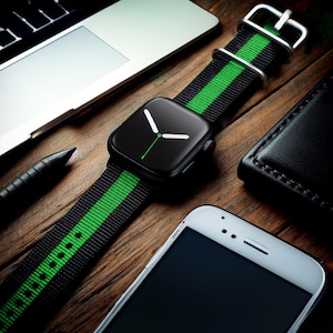 May include: A black and green striped watch band with a black Apple Watch face. The watch is on a wooden surface with a laptop, a pen, and a black leather wallet.