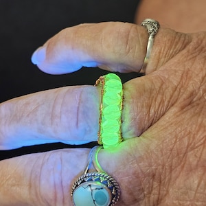 May include: A ring featuring a series of small, round, neon green beads arranged in a row and secured with thin, metallic copper-colored wire. The beads emit a bright, glowing light, contrasting with the dark background and metallic wire structure.