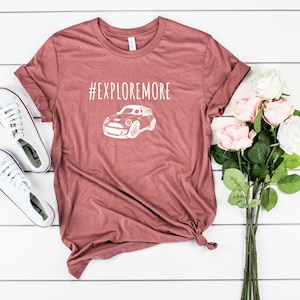 Puede incluir: Una camiseta de color marrón rojizo con un gráfico blanco de un coche y el texto "#EXPLOREMORE".