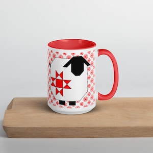 May include: A white ceramic mug with a red interior and handle. The mug features a sheep design with a black head and a red star on its body. The background has a repeating red floral pattern. The mug is placed on a wooden surface.