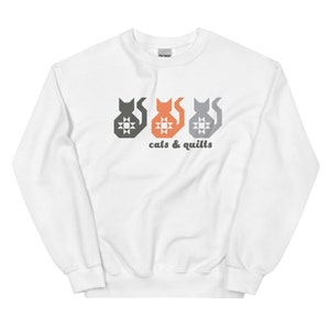 May include: White sweatshirt featuring a graphic of three stylized cat silhouettes in gray, orange, and gray, each with a starburst design. The text "cats & quilts" is printed below the cats in a dark gray font. The sweatshirt is made of a soft material.