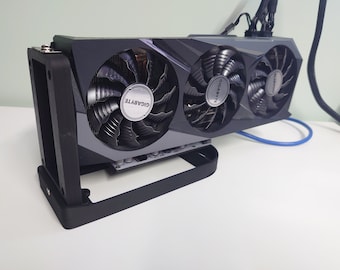 May include: A black graphics card with three fans, each with a black and silver design and the word "GIGABYTE" printed on them. The card is mounted on a black stand.