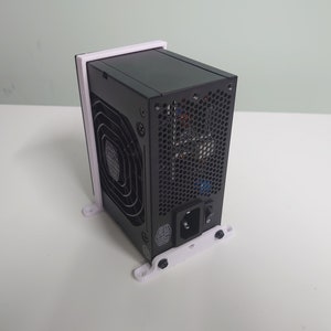 K&ouml;nnte beinhalten: Ein schwarzes Netzteil f&uuml;r Computer, das auf einer wei&szlig;en, 3D-gedruckten Halterung montiert ist. Das Netzteil hat einen L&uuml;fter und ein Netzkabel.
