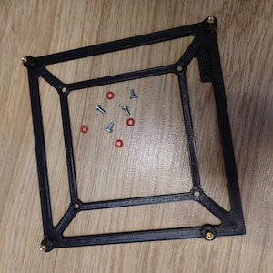 May include: A black, 3D-printed square frame with brass-coloured corner accents. Several small screws and red O-rings are scattered inside the frame. The frame is resting on a light brown wooden surface.