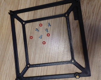 May include: A black, 3D-printed square frame with brass-colored corner accents. Several small screws and red O-rings are scattered inside the frame. The frame is resting on a light brown wooden surface.