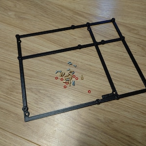 Puede incluir: Marco de plástico negro con tornillos y pequeños herrajes rojos y dorados. El marco es rectangular con varias líneas que se cruzan y esquinas redondeadas. Los herrajes incluyen tornillos pequeños, arandelas y otros componentes.