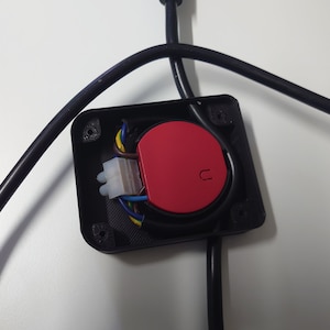 Pode incluir: Close-up de uma caixa elétrica preta com um botão vermelho e um conector branco. A caixa está conectada a fios pretos.