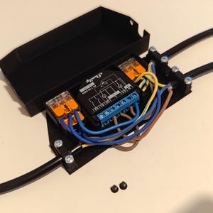 May include: A black plastic electrical box with a clear lid, containing a black electrical device with blue and orange wiring. The device has the text "Shelly 2.5" printed on it. The box is mounted on a white surface.