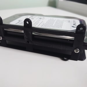 Puede incluir: Una carcasa de disco duro negra con un disco duro plateado en el interior. La carcasa tiene un marco rectangular con orificios para tornillos y está diseñada para sujetar el disco duro de forma segura. El disco duro es visible a través del marco.