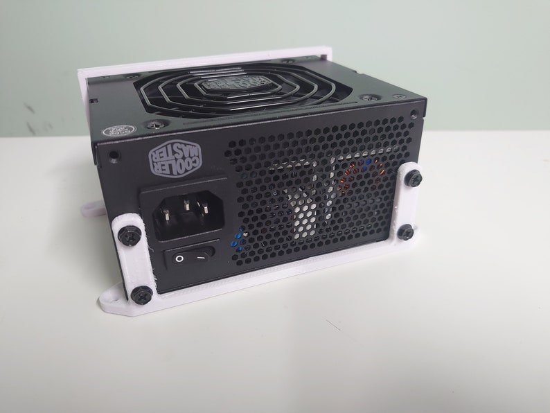 K&ouml;nnte beinhalten: Ein schwarzes Netzteil von Cooler Master mit einer wei&szlig;en Kunststoffhalterung. Das Netzteil hat einen L&uuml;fter, einen Netzschalter und einen Netzstecker.