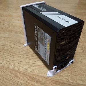 Może przedstawiać: Czarno-biały zasilacz Corsair RM750i z białym wspornikiem. Zasilacz ma czarną siatkę i białą etykietę z logo Corsair i tekstem "RM750i".