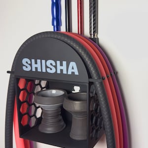Könnte beinhalten: Schwarzer, wandmontierter Shisha-Ständer mit einem Regal für Schalen und einem Haken für Schläuche. Der Ständer trägt die Aufschrift "SHISHA" auf der Vorderseite. Vier Schläuche hängen an den Haken, zwei rote, ein lila und ein schwarzer.