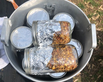 May include: A silver metal bucket filled with glass jars of preserved food. The jars are wrapped in aluminum foil and contain a mixture of grains and seeds.