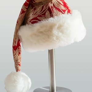 Könnte beinhalten: Ein festlicher Weihnachtsmannhut aus rotem Stoff mit goldfarbener Blattstickerei. Der Hut hat einen flauschigen weißen Kunstfellbesatz und einen passenden Bommel. Ideal für Weihnachtsdekorationen.