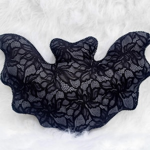 Peut inclure: Un coussin en forme de chauve-souris en dentelle noire avec un motif floral détaillé. Le coussin est posé sur un fond blanc et duveteux. Ce coussin décoratif est parfait pour Halloween ou une décoration gothique.