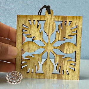 May include: A square wooden ornament with a snowflake design cut out. The ornament is held by a black string and has a natural wood grain finish. The background is a light blue.