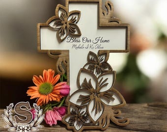 Op de afbeelding: Een houten kruis met een witte achtergrond en bruine bloemmotieven. Het kruis heeft de tekst "Bless Our Home Mahalo I Ke Akua" erop.