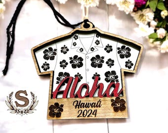 May include: Wooden ornament shaped like a Hawaiian shirt with a black and white floral pattern. The ornament has the word "Aloha" in red lettering and the text "Hawaii 2024" below it.