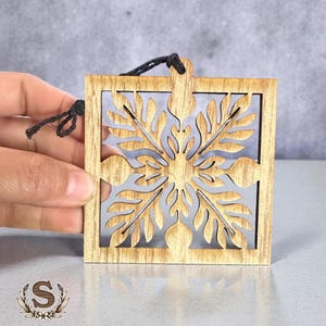 May include: A square wooden ornament with a floral design cut out of the center. The ornament is light brown with a black string for hanging. The design is symmetrical and detailed, perfect for holiday decor.