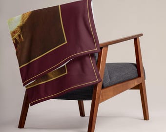 Puede incluir: Una manta rectangular colocada sobre un sillón de madera. La manta presenta un borde de color burdeos oscuro con finas líneas doradas que enmarcan una imagen central de una figura de cabello dorado con corona, sobre un fondo marrón oscuro.