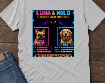 Puede incluir: Una camiseta gris claro con un gráfico de estilo arcade de 16 bits titulado LUNA & MILO SELECT YOUR FIGHTER. El diseño muestra un bulldog francés llamado Luna y un golden retriever llamado Milo con barras de salud y velocidad debajo.
