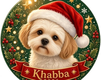 Puede incluir: Un adorno navideño redondo con un cachorro blanco y beige que lleva un gorro de Papá Noel rojo. El fondo es verde oscuro con acebo, bayas rojas y estrellas doradas. Una cinta roja en la parte inferior muestra el texto Khabba 2026.