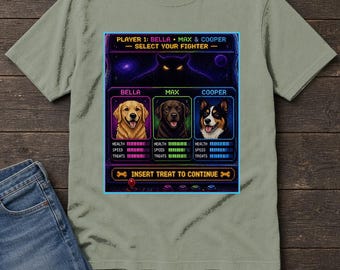 Puede incluir: Una camiseta verde salvia con un gráfico de juego arcade de 16 bits. El diseño muestra tres perros llamados Bella, Max y Cooper con estadísticas de salud, velocidad y premios. El texto incluye PLAYER 1: BELLA, MAX & COOPER, SELECT YOUR FIGHTER y INSERT TREAT TO CONTINUE.