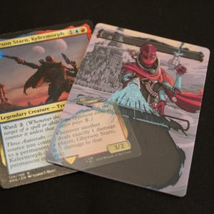 Könnte beinhalten: Zwei Magic: The Gathering Sammelkarten. Eine Karte zeigt eine Figur mit einer Waffe in einer Wüstenlandschaft. Die andere Karte zeigt eine Figur in Rot und Blau mit einer Waffe vor einem schneebedeckten Hintergrund. Die Karten haben Text.