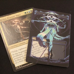 May include: Two Magic: The Gathering trading cards. One card features a gold and tan illustration of a creature with the text "Atraxa, Praetors' Voice". The other card has a dark background with a teal and white illustration of a humanoid figure.