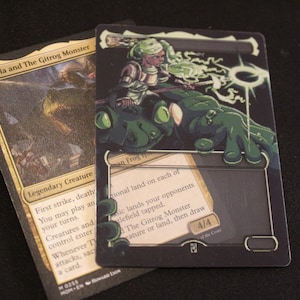 Puede incluir: Dos cartas coleccionables. Una carta presenta el texto "Thalia and The Gitrog Monster" con un borde dorado y una ilustración de criatura. La otra carta tiene un fondo oscuro con una ilustración verde y blanca de un personaje montando una criatura parecida a una rana.