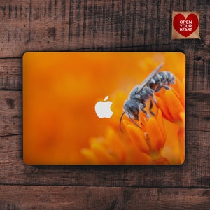 Könnte beinhalten: Nahaufnahme einer Biene auf einer leuchtend orangefarbenen Blüte. Die Biene ist schwarz und grau mit einem weißen Apple-Logo im Hintergrund. Das Bild ist auf einem Laptop.