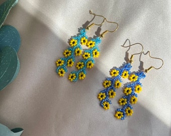 Puede incluir: Un par de pendientes colgantes con un diseño floral. Los pendientes están hechos de cuentas azules y amarillas y tienen ganchos dorados.