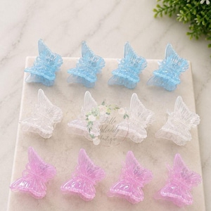 May include: A collection of twelve butterfly-shaped hair clips arranged in three rows of four. The top row features four light blue glittery clips, the middle row has four clear clips, and the bottom row contains four translucent pink butterfly clips.