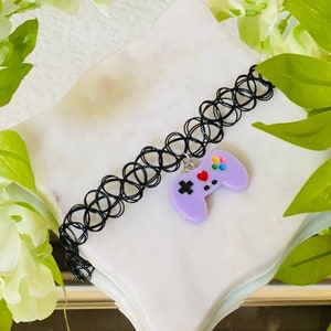May include: A black choker necklace with a purple video game controller charm. The controller has colorful buttons and a red heart on the front.
