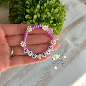 Puede incluir: Un brazalete de cuentas moradas y blancas con cuentas de margarita blancas y la palabra "BIG SIS" escrita en letras negras. Un pequeño dije en forma de corazón está en el centro del brazalete.