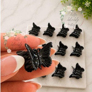 May include: A collection of small, glossy black butterfly-shaped hair clips. One clip is held in the foreground, while eight identical clips are arranged in a grid pattern on a square white tile. Each clip features detailed wing textures.
