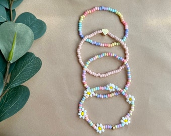 May include: Four colorful beaded bracelets with a daisy charm on the bottom bracelet. The bracelets are stacked on top of each other and are on a light brown background.