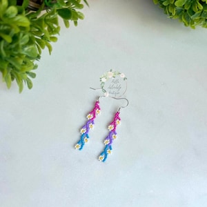 May include: A pair of dangle earrings with a gradient of pink, purple, blue, and white beads. The beads are arranged in a flower shape. The earrings are made with silver hooks.