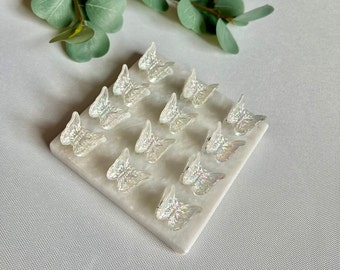 May include: A set of 12 iridescent butterfly hair clips. The clips are arranged in a grid pattern on a white surface. The clips are made of plastic and have a shiny, iridescent finish.