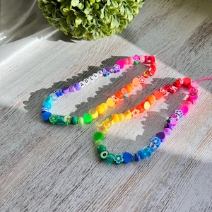May include: A colorful beaded necklace with the name "ADELE" spelled out in white letters. The beads are arranged in a rainbow pattern and include various shapes, such as flowers, hearts, and stars.