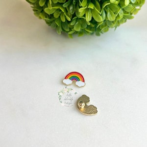 May include: A rainbow enamel pin with white clouds and a gold backing. The pin features a vibrant rainbow design with red, orange, yellow, green, blue, and purple colours. The pin is small and detailed.