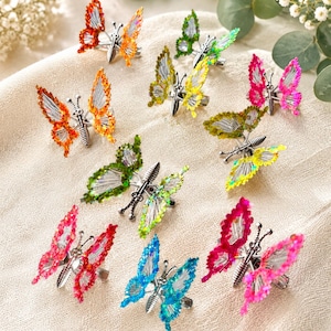 May include: A collection of ten butterfly-shaped hair clips featuring silver-toned metal bodies and translucent, vibrant wings in shades of orange, yellow, green, blue, pink, and red. Each clip has a textured, sparkling finish on the wing edges.