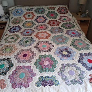 May include: A handmade quilt featuring a repeating pattern of floral fabric hexagons arranged in flower-like clusters. The quilt has a cream-coloured background with various multi-coloured printed cotton patches in shades of pink, blue, green, and red, finished with a thin floral border along the bottom edge.