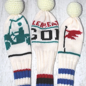 Puede incluir: Tres fundas para palos de golf. Cada una es de color crema con un diseño de punto. Una tiene un patrón verde y blanco, otra tiene la palabra "LEPER" y el número "301", y la tercera tiene una bandera roja. Las bases tienen rayas.